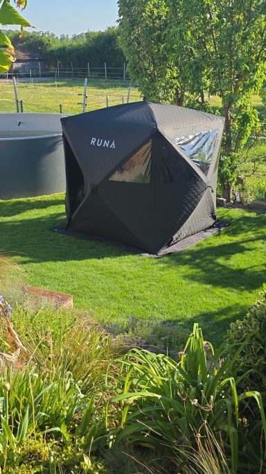 Geometrisches, schwarzes Gartenhaus auf grünem Rasen, umgeben von Bäumen und Sträuchern.