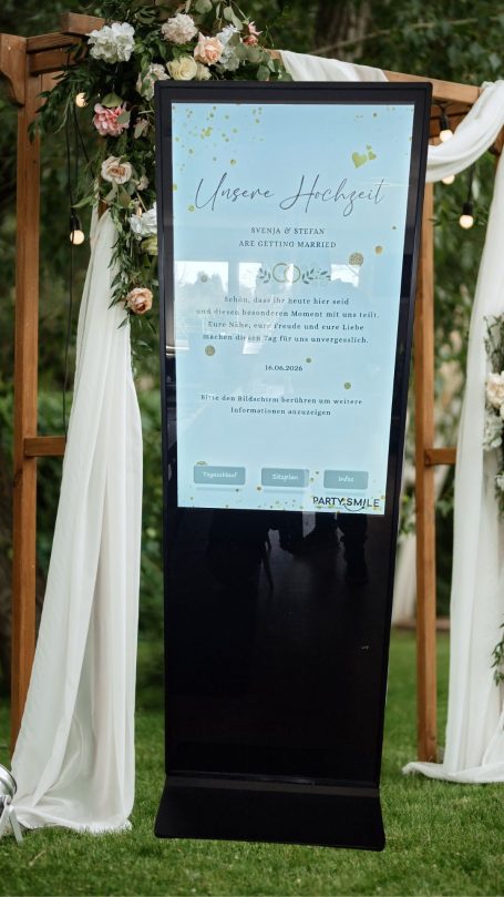 Tafel im Freien mit Veranstaltungshinweisen, umgeben von Blumen und Stoffvorhängen.