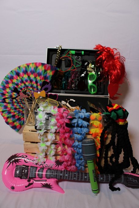 Bunte Partyutensilien, darunter Gitarre, Mikrofon und Perücken, in einer Kiste arrangiert.