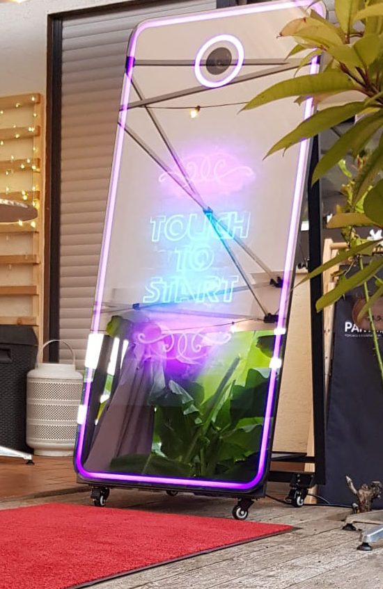 Magic Mirror Ein moderner Fotobox-Spiegel mit bunten Lichtern und Pflanzen im Hintergrund.