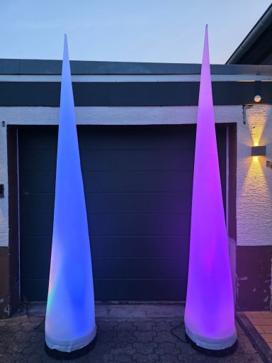Zwei beleuchtete, konische Skulpturen in Blau und Lila vor einer Garagenauffahrt.