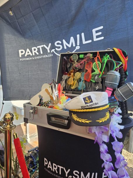 Ein Koffer voller Party-Accessoires wie Hüte, Brillen und Mikrofone, vor einem Banner.